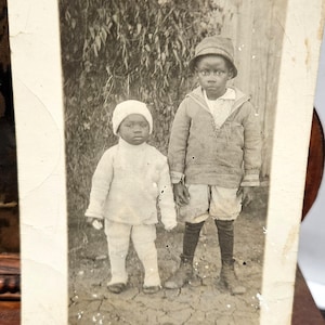 May include: Vintage black and white photograph featuring two young children. One child wears a white knit cap, sweater, and pants. The other child is in a hat, jacket, striped shirt, shorts, and long socks. The photo is aged and shows signs of wear.