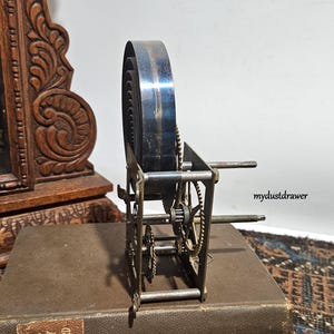 May include: An antique clock spring mechanism with visible gears and a large, coiled spring. The metal components have a vintage appearance, suggesting age and use. The mechanism is set on a brown book.