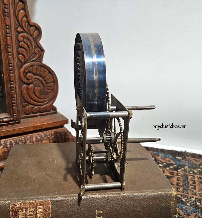 May include: An antique clock spring mechanism with visible gears and a large, coiled spring. The metal components have a vintage appearance, suggesting age and use. The mechanism is set on a brown book.