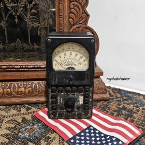 May include: A vintage black Supreme Model 542 multimeter with a circular gauge and multiple dials. The device sits on a patterned rug next to a small American flag. The background includes a wooden decorative piece.