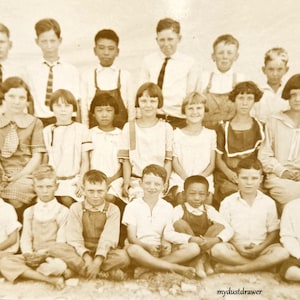May include: Sepia-toned vintage photograph of a group of children and young adults, likely a school class. The subjects are arranged in rows, with some seated and others standing. Most are wearing light-colored shirts and ties.