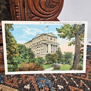 May include: Vintage postcard featuring a color illustration of the South Approach to Sanitarium in Battle Creek, Michigan. The image shows a large building with a flag, surrounded by trees and a grassy lawn. The postcard is set against a wooden background.