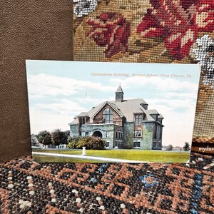 May include: A vintage postcard depicting the Gymnasium Building of the Normal School in West Chester, Pennsylvania. The building is a large, stone structure with a central tower and a gabled roof. The postcard is in color and shows the building against a blue sky with white clouds.