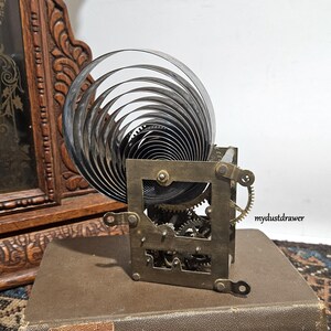 May include: A vintage clock movement with a large, coiled spring and intricate gears. The metal components are aged, with a square frame and small brass gears. The clock mechanism sits on a brown surface, suggesting a historical piece.