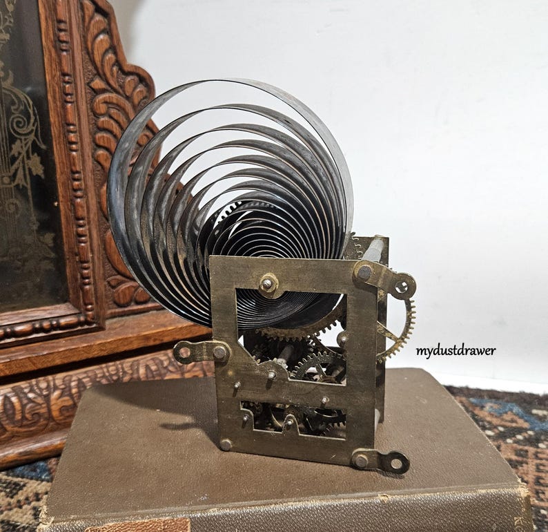 May include: A vintage clock movement with a large, coiled spring and intricate gears. The metal components are aged, with a square frame and small brass gears. The clock mechanism sits on a brown surface, suggesting a historical piece.