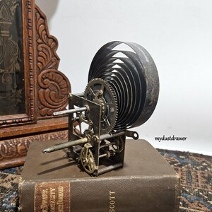 May include: An antique clock movement with visible gears and a large coiled spring. The metal components are aged, with a dark patina. The clock mechanism sits on a vintage book, with a carved wooden clock in the background.