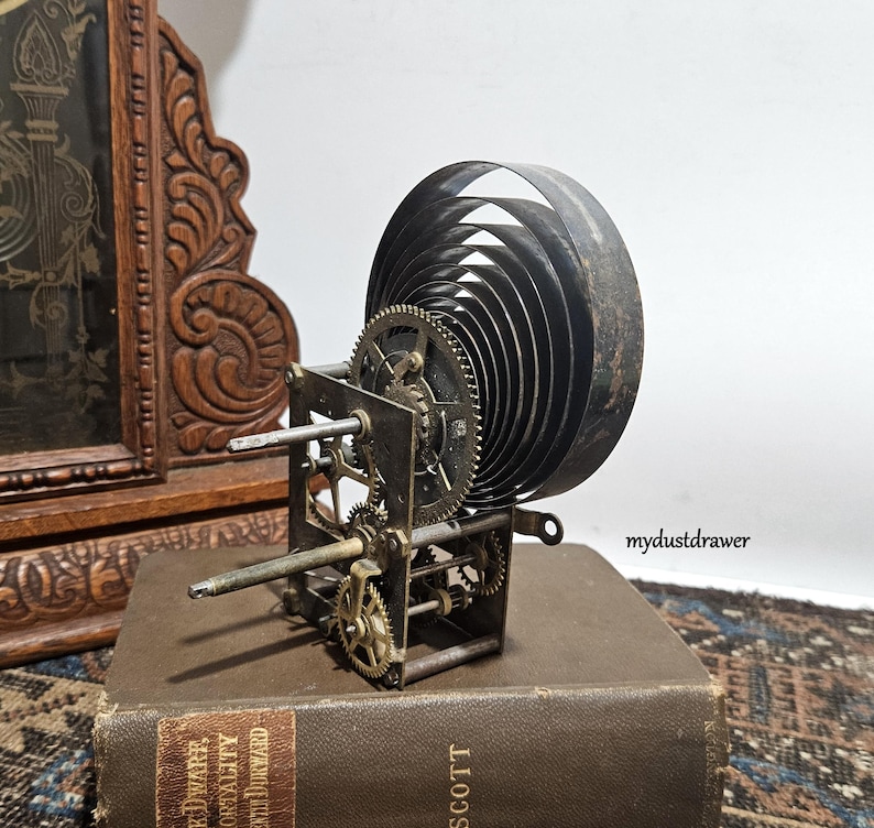 May include: An antique clock movement with visible gears and a large coiled spring. The metal components are aged, with a dark patina. The clock mechanism sits on a vintage book, with a carved wooden clock in the background.