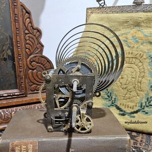 May include: A vintage clock movement with visible gears and a curved metal spring. The mechanism is set on a brown book, with a carved wooden frame and a tapestry bag in the background. The clock mechanism is made of metal.