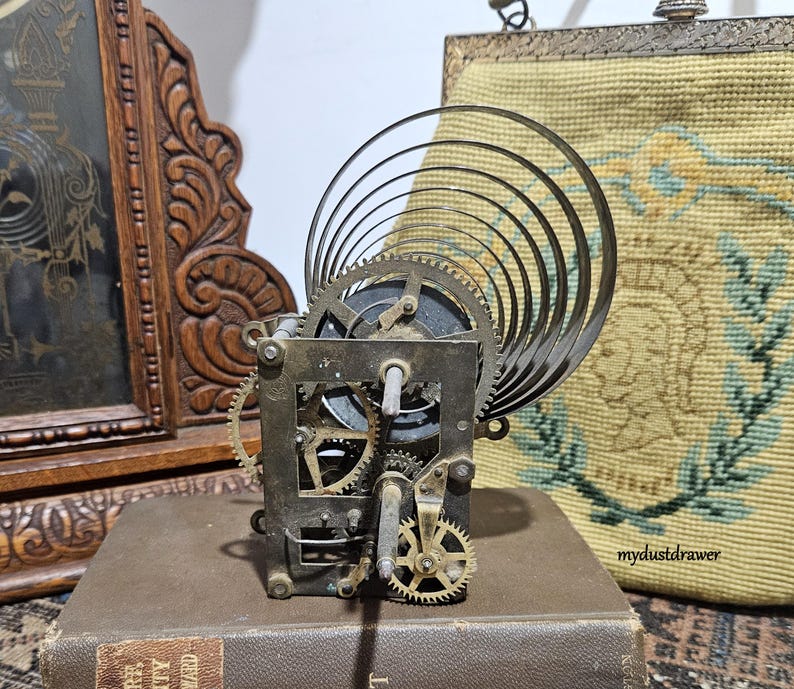 May include: A vintage clock movement with visible gears and a curved metal spring. The mechanism is set on a brown book, with a carved wooden frame and a tapestry bag in the background. The clock mechanism is made of metal.