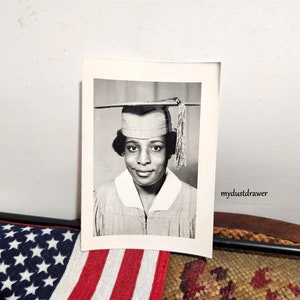 May include: Black and white photograph of a woman wearing a graduation cap and gown. She is smiling and looking at the camera. The photo is in a white frame.