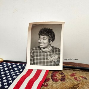 May include: Black and white photograph of a woman with short, curly hair wearing a plaid shirt and a bow. The photo is in a white frame and has the text "mydustdrawer" in the bottom right corner.