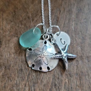 May include: A silver necklace with a sea-themed charm set. The charms include a sand dollar, a starfish, and a light blue glass bead. The necklace also has a silver charm with the letter "C" on it.