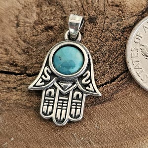 May include: A silver-colored Hamsa hand pendant with a turquoise stone center. The pendant is next to a dime coin on a wooden surface. The Hamsa hand is a symbol of protection and good fortune.