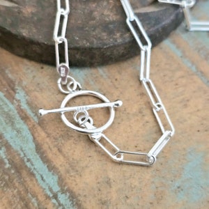 May include: Silver chain necklace with a toggle clasp. The chain is made of linked oval shapes.
