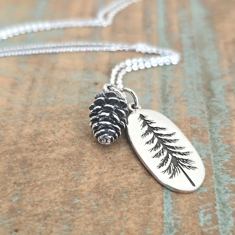 May include: A silver necklace with a pine tree charm and a pine cone charm. The charms are both detailed and realistic.