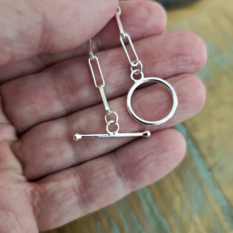 May include: Silver chain with a toggle clasp and a circular ring. The chain is made of small, rectangular links.