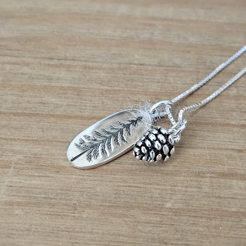May include: A silver necklace with a pine tree charm and a pine cone charm. The necklace is on a wooden surface.