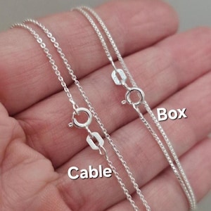 May include: Two silver chains, one labeled "Cable" and the other labeled "Box", are shown on a hand. The chains are both silver and have a clasp at the end.