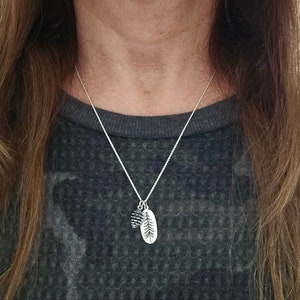 May include: A silver chain necklace with a small pine cone charm and a larger oval charm with a pine tree design.