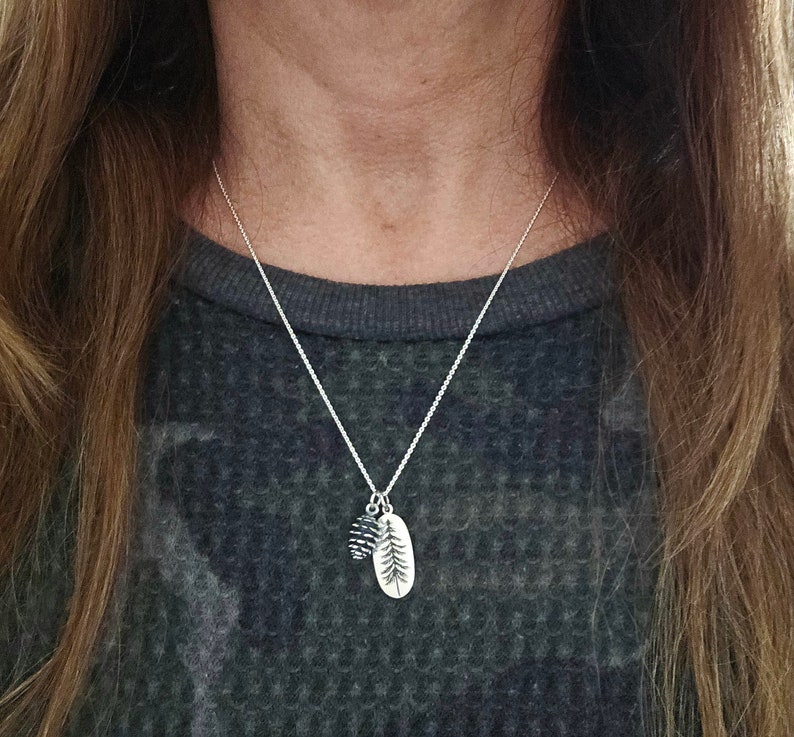 May include: A silver chain necklace with a small pine cone charm and a larger oval charm with a pine tree design.