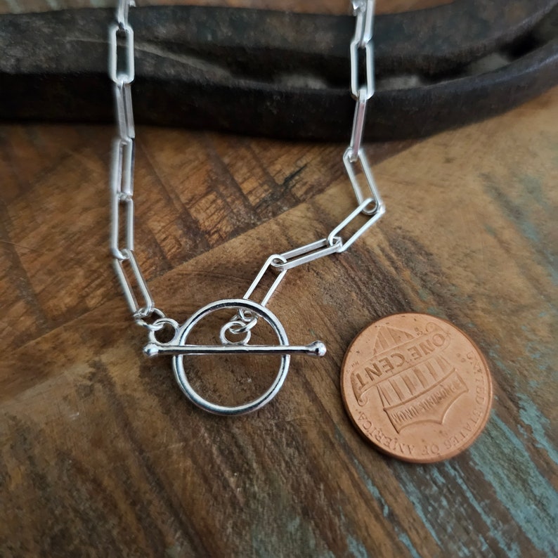 May include: Silver chain necklace with a toggle clasp. The chain is made of flat, rectangular links. The toggle clasp is a round ring with a bar that slides through it. The necklace is shown next to a US penny for scale.