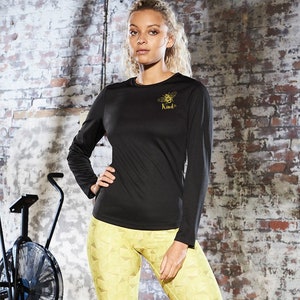 May include: Black long-sleeve shirt with a gold bee and the word "Kind" on the front. The shirt is worn by a person with long blonde hair.