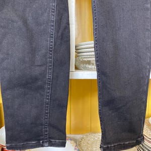 Pu&ograve; includere: Un paio di jeans in denim nero a gamba dritta. I jeans sono appesi a uno scaffale con ripiani bianchi e pareti gialle.
