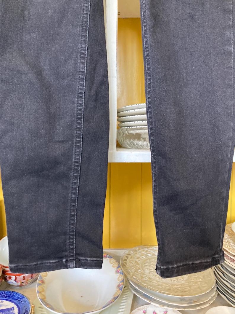 Pu&ograve; includere: Un paio di jeans in denim nero a gamba dritta. I jeans sono appesi a uno scaffale con ripiani bianchi e pareti gialle.