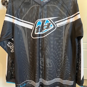 Könnte beinhalten: Schwarzes Mesh-Trikot mit weißen und blauen Akzenten. Das Trikot zeigt das Logo "Troy Lee Designs" am Kragen und ein großes Logo auf der Brust. Weiße Streifen verlaufen über den Schultern und Ärmeln. Das Trikot hat einen V-Ausschnitt und lange Ärmel.