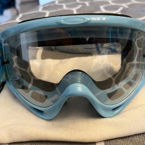 May include: Light blue Oakley goggles with a clear lens. The goggles have a black strap and a blue and white patterned strap. The Oakley logo is visible on the side. The goggles are sitting on a white cloth.