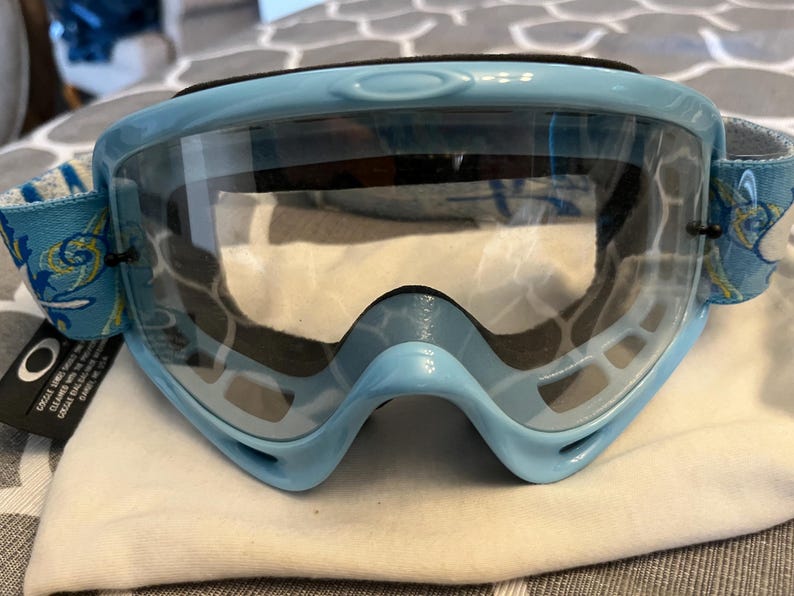 May include: Light blue Oakley goggles with a clear lens. The goggles have a black strap and a blue and white patterned strap. The Oakley logo is visible on the side. The goggles are sitting on a white cloth.