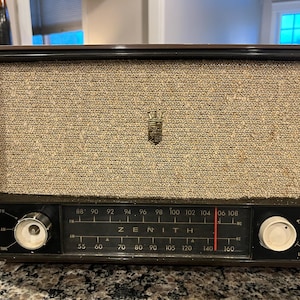 Może przedstawiać: Zabytkowe radio Zenith z brązową drewnianą ramą i beżową osłoną głośnika z tkaniny. Radio posiada dwa pokrętła, pokrętło częstotliwości i logo Zenith. Tarcza wyświetla częstotliwości AM i FM.