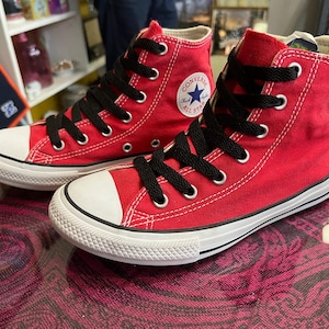May include: Pair of red high-top canvas sneakers with black laces and white rubber soles. The sneakers feature the Converse All Star logo on the side. The shoes are new and unworn.