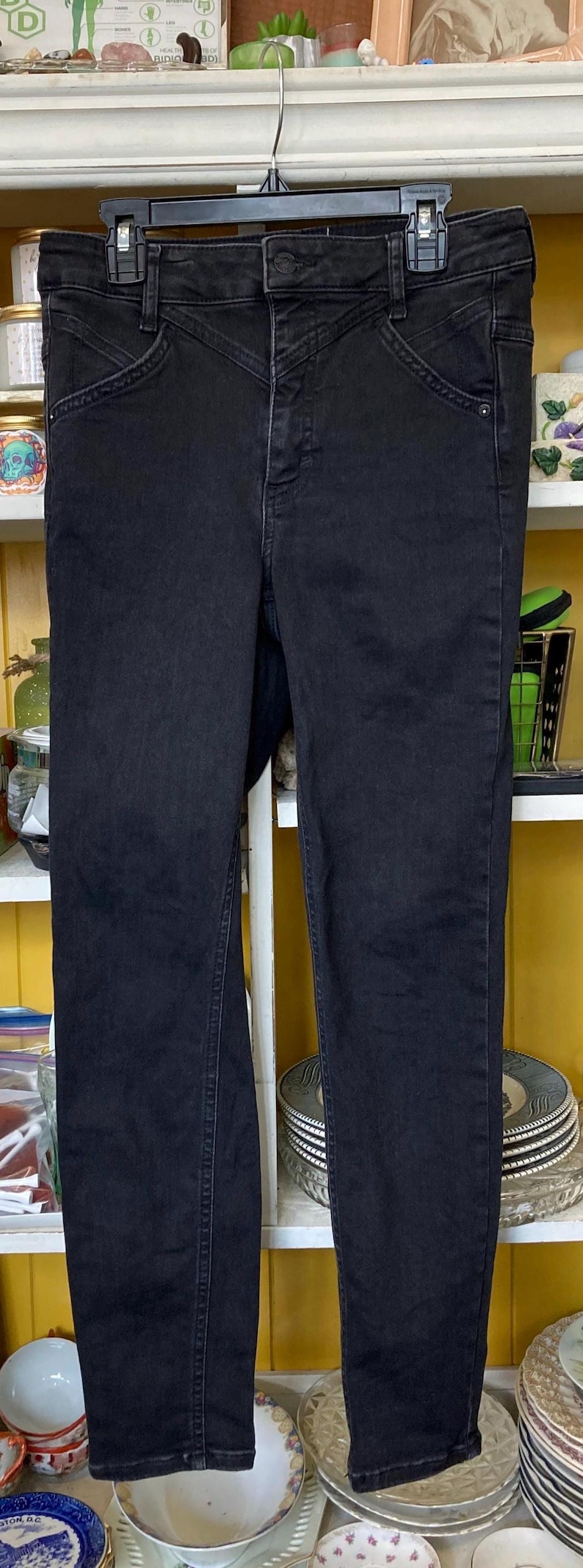Pu&ograve; includere: Jeans skinny neri a vita alta con zip. I jeans hanno un aspetto leggermente rovinato e sono appesi a un'appendiabiti nero.