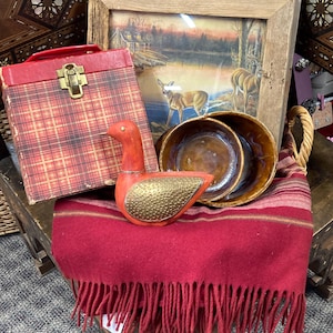 Puede incluir: Una exhibición vintage con una caja a cuadros roja, una pintura de paisaje enmarcada con ciervos y dos cuencos marrones. Una figura de ganso rojo con un ala dorada texturizada se encuentra sobre una manta roja con flecos. Los artículos están dispuestos sobre un cofre de madera.