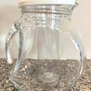 May include: A clear glass jug with a white lid and a clear plastic ice tube insert. The jug has a curved handle and a wide base. The jug is on a speckled countertop.