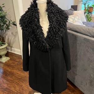 May include: A black coat with a textured, dark-colored collar. The coat is displayed on a white mannequin. The coat has long sleeves and a tailored fit. The background includes a wooden floor and a gray sofa.