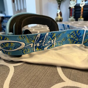 May include: A pair of Oakley ski goggles with a light blue frame and a black padded interior. The strap is light blue with a floral design and the Oakley logo in white. The goggles rest on a white cloth.