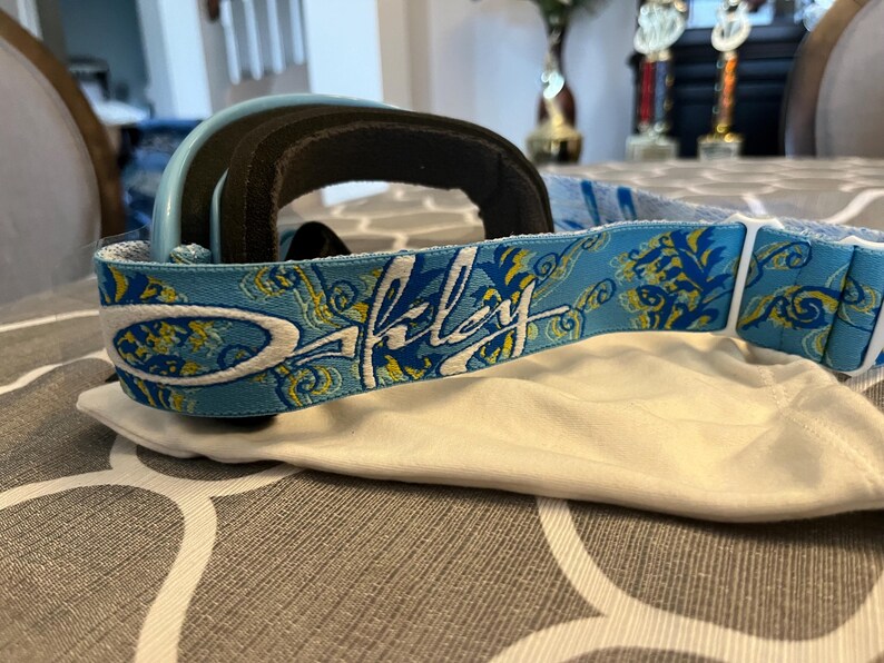 May include: A pair of Oakley ski goggles with a light blue frame and a black padded interior. The strap is light blue with a floral design and the Oakley logo in white. The goggles rest on a white cloth.