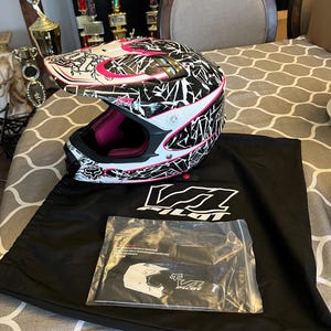 Könnte beinhalten: Ein weißer, schwarzer und pinkfarbener Motocross-Helm mit geometrischem Design. Der Helm hat eine rosa Innenausstattung und liegt neben einer schwarzen Helmtasche mit dem Logo "V1 Pilot" und einer transparenten Plastiktüte mit Anweisungen.