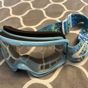 May include: Light blue ski goggles with a black foam lining and a patterned strap. The goggles have a clear lens and a light blue frame. The strap features a blue and yellow floral design.