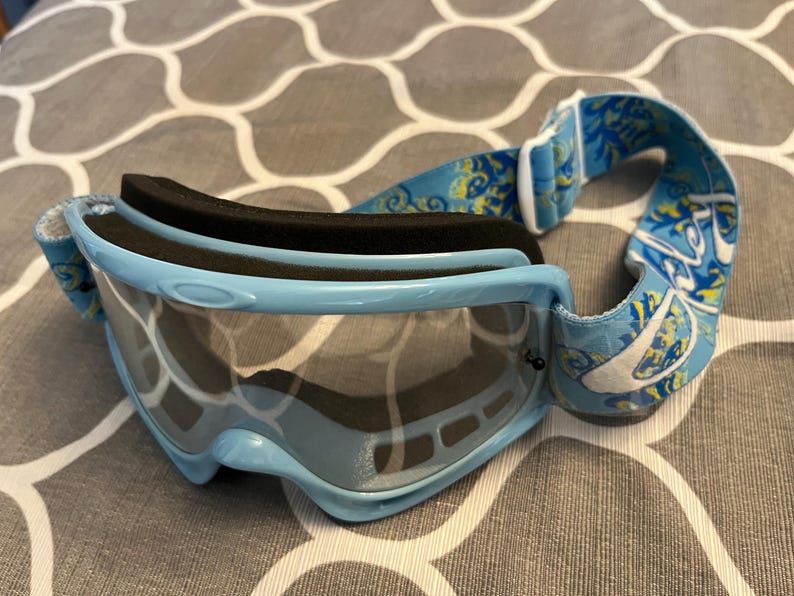 May include: Light blue ski goggles with a black foam lining and a patterned strap. The goggles have a clear lens and a light blue frame. The strap features a blue and yellow floral design.