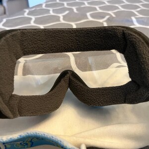 May include: Close-up of ski goggles with a black, textured frame and clear lenses. The interior features a thick, dark gray padding for comfort. The strap has a blue and yellow floral pattern.