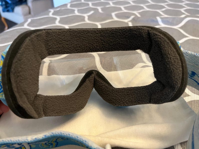 May include: Close-up of ski goggles with a black, textured frame and clear lenses. The interior features a thick, dark gray padding for comfort. The strap has a blue and yellow floral pattern.
