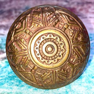 May include: A detailed, antique brass doorknob with an intricate, embossed design. The central circle features a floral pattern, surrounded by geometric shapes and lines. The knob has a warm, aged patina, suggesting it is vintage.