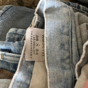 以下が含まれることがあります： 明るいブルーデニムのジーンズのクローズアップ。ジーンズは色あせたウォッシュ加工と目立つステッチが施されています。サイズ38 x 34などの詳細が記載されたタグが見えます。ジーンズは折りたたまれており、内側の裏地とポケットが見えています。