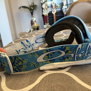 May include: Blue and black ski goggles with a patterned strap. The strap features a light blue background with a floral design in yellow and white. The brand name "Oakley" is prominently displayed in white.