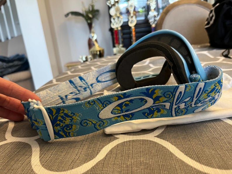 May include: Blue and black ski goggles with a patterned strap. The strap features a light blue background with a floral design in yellow and white. The brand name "Oakley" is prominently displayed in white.