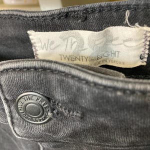 Pu&ograve; includere: Primo piano di un paio di jeans neri con un'etichetta We The Free Twenty Eight e un bottone nero.