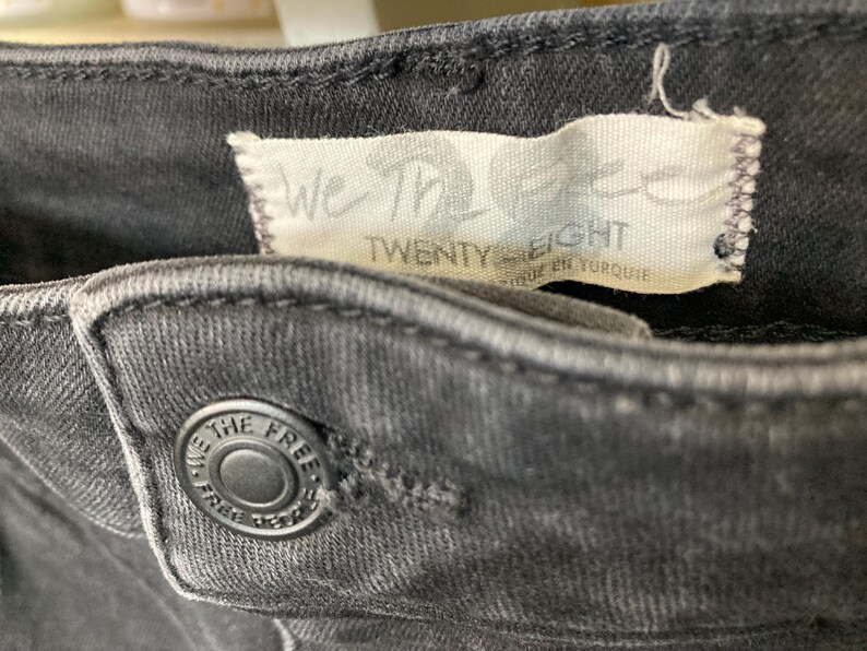 Pu&ograve; includere: Primo piano di un paio di jeans neri con un'etichetta We The Free Twenty Eight e un bottone nero.